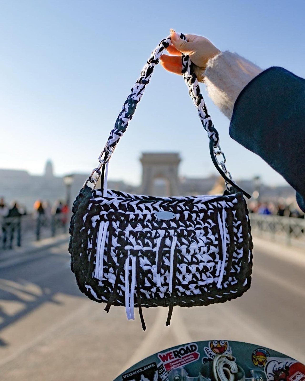 A black-and-white crochet Meraki bag held up by a river in Budapest