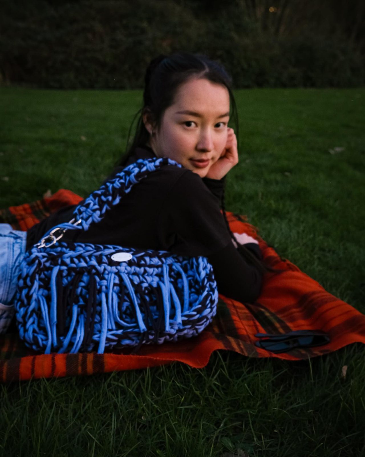 A woman lying on a picnic blanket with a blue crochet Meraki bag